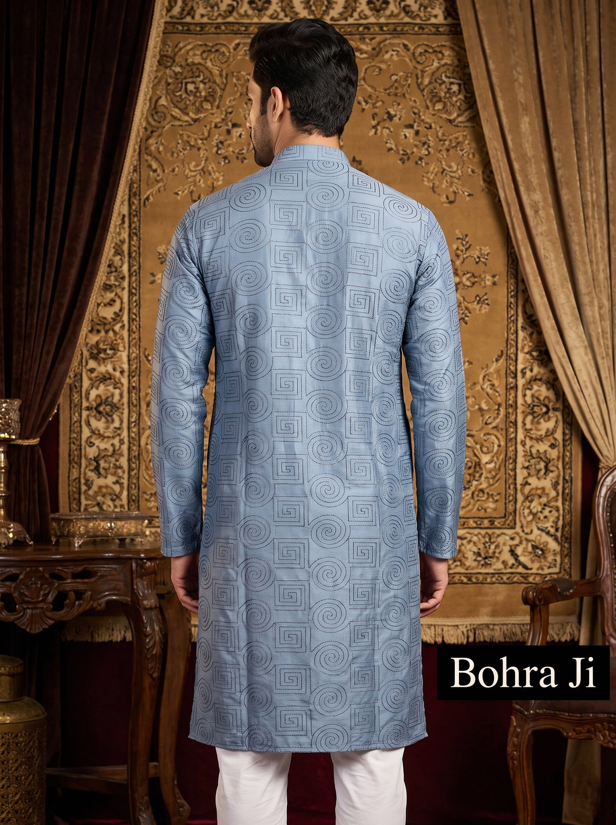 A man viewed from the back wearing a light blue patterned kurta with white trousers, standing in front of an ornate gold patterned tapestry. The brand name 'Bohra Ji' is displayed in a black box in the bottom right corner.