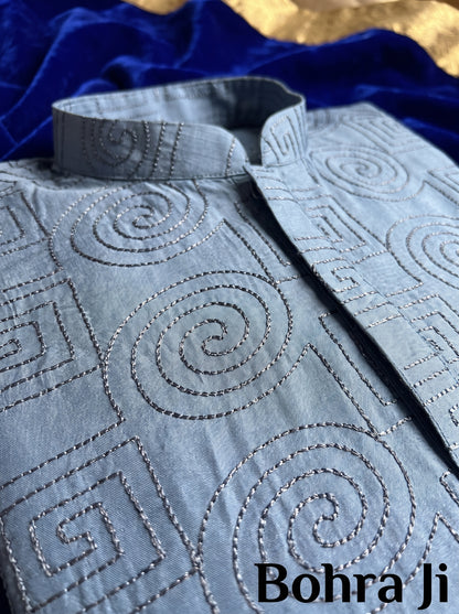 A close-up of a light blue embroidered kurta fabric with spiral and geometric stitched patterns, draped over a dark blue velvet surface with a gold fabric accent in the background. The text 'Bohra Ji' is visible in the bottom right corner.
