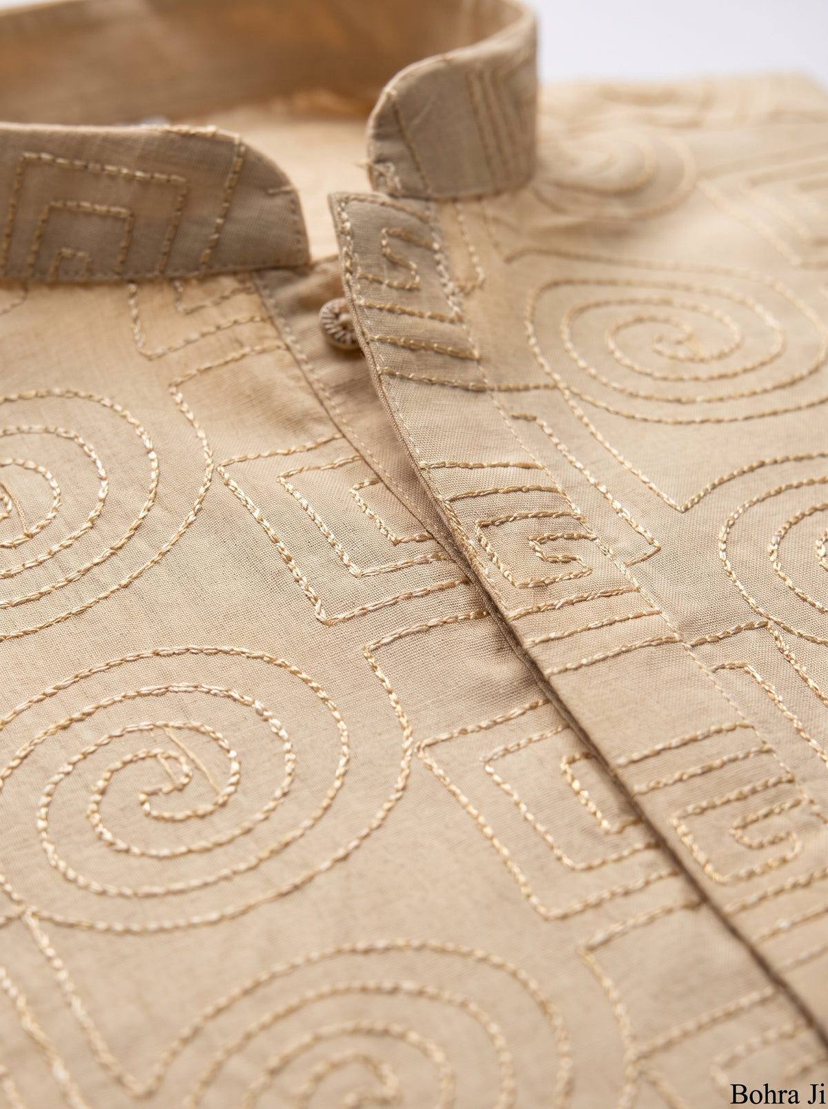 Close-up of a beige fabric garment with tonal geometric and spiral embroidery, showing the collar and front placket.
