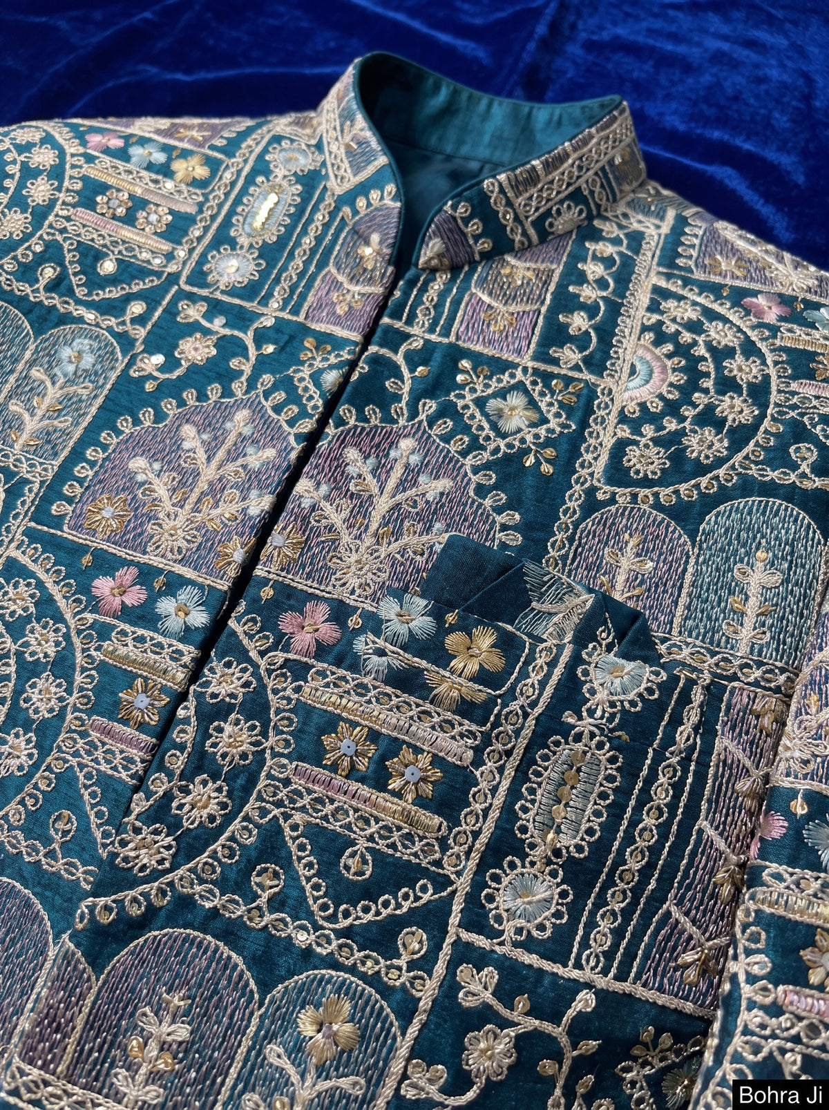 A close-up of an ornate teal blue embroidered jacket or sherwani with intricate gold, beige, and purple paisley and floral patterns, featuring a mandarin collar and a chest pocket.