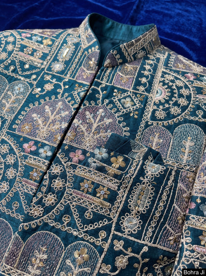 A close-up of an ornate teal blue embroidered jacket or sherwani with intricate gold, beige, and purple paisley and floral patterns, featuring a mandarin collar and a chest pocket.