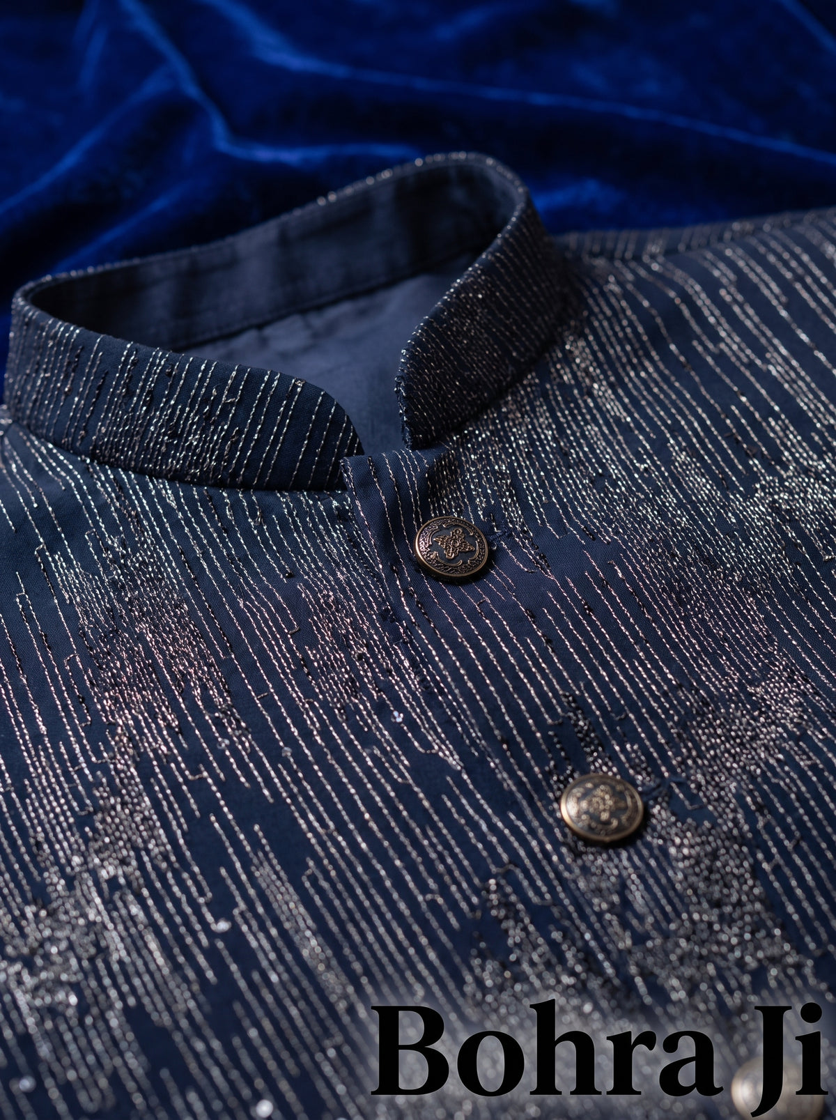 A close-up of a dark navy blue traditional sherwani jacket with shimmering silver metallic vertical stripes and bronze buttons, set against a dark blue velvet background. The brand name 'Bohra Ji' is visible as a watermark at the bottom right.