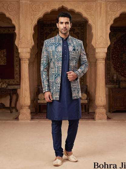 A man modeling a traditional Indian outfit consisting of a navy blue kurta and trousers paired with a blue and white patterned sherwani jacket, standing in front of an ornate stone archway.