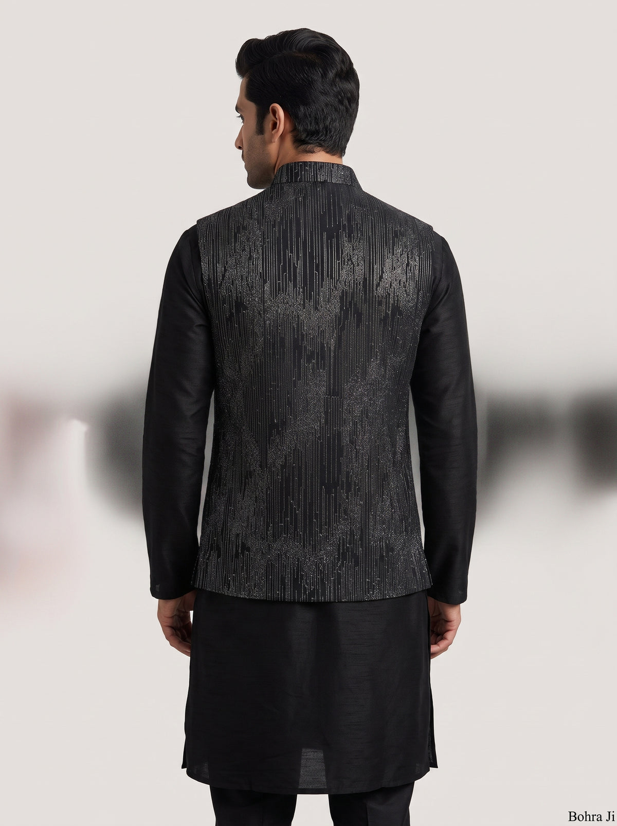 A man viewed from the back wearing a black long kurta with a textured, vertically patterned waist jacket. The jacket features a subtle metallic sheen with vertical stripes of black, gray, and silver.