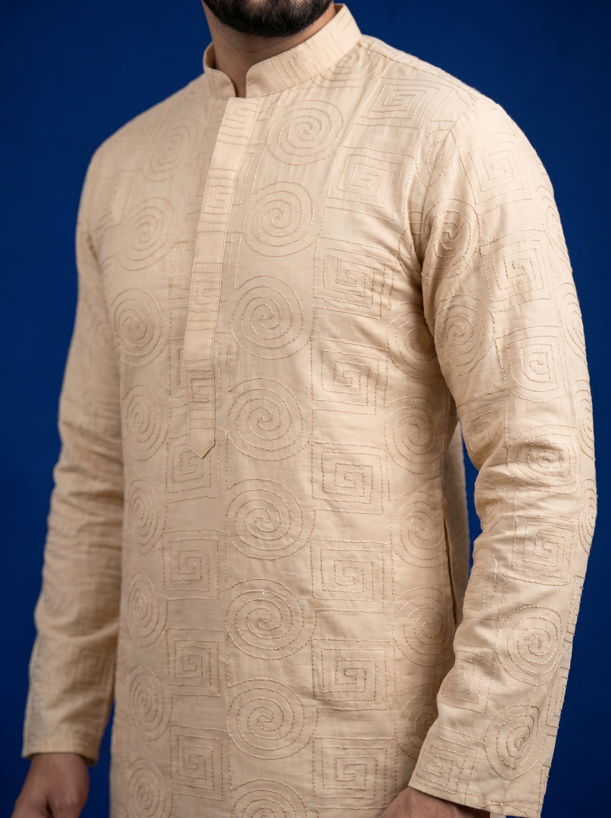 A man wearing a beige long-sleeve kurta with an allover tonal geometric and spiral jacquard pattern, shown against a solid dark blue background.