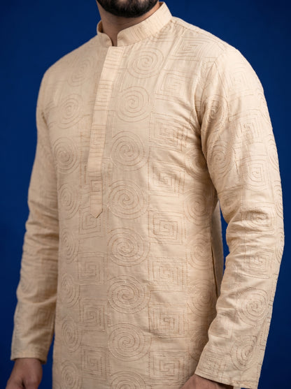 A man wearing a beige long-sleeve kurta with an allover tonal geometric and spiral jacquard pattern, shown against a solid dark blue background.