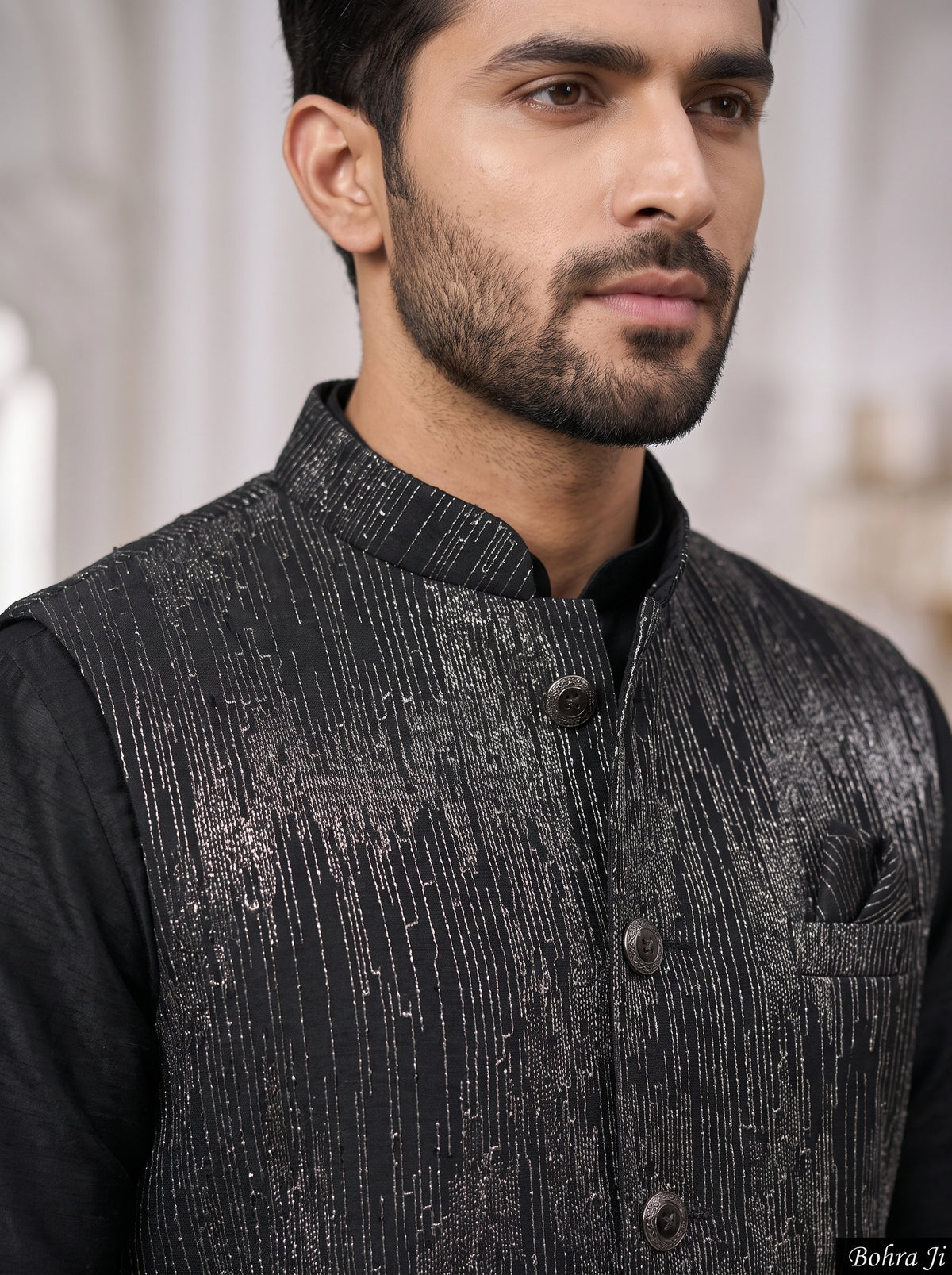 A close-up of a man wearing a black textured waistcoat with vertical metallic-looking stripes over a black shirt. The background is a softly blurred indoor setting.