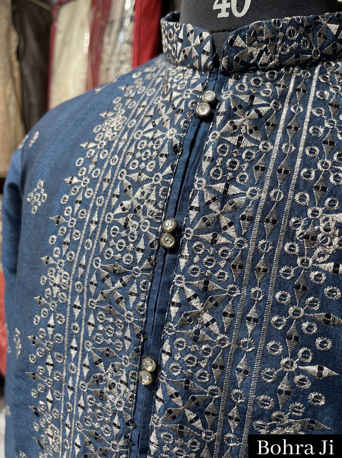 A close-up of a blue embroidered kurta or shirt on a mannequin, featuring intricate silver and white geometric patterns, a high collar, and decorative buttons. The text 'Bohra Ji' is visible in the bottom right corner.