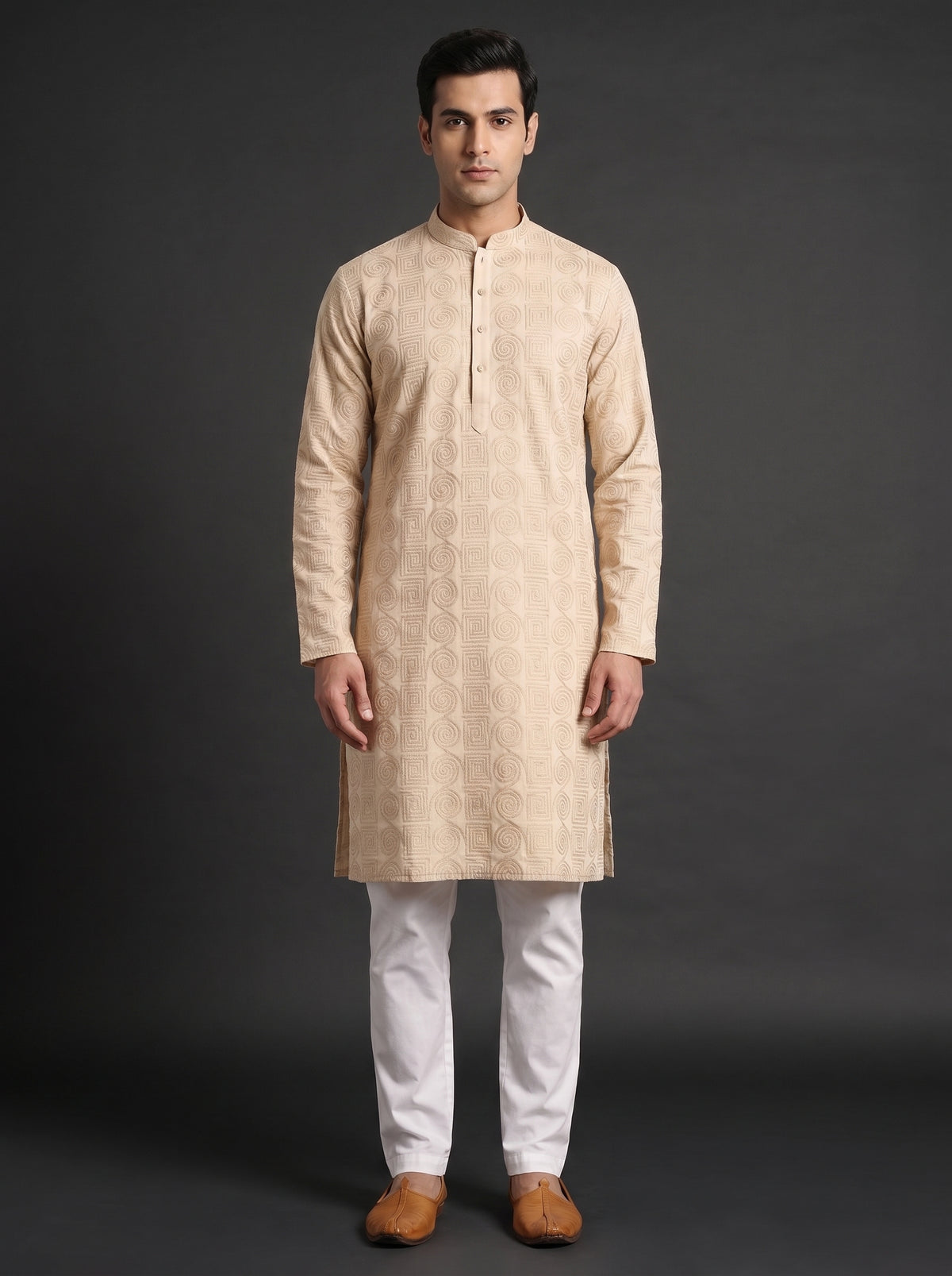 A man modeling a light beige kurta with subtle gold-toned embroidery, paired with white trousers and brown shoes, standing against a dark gray studio background.