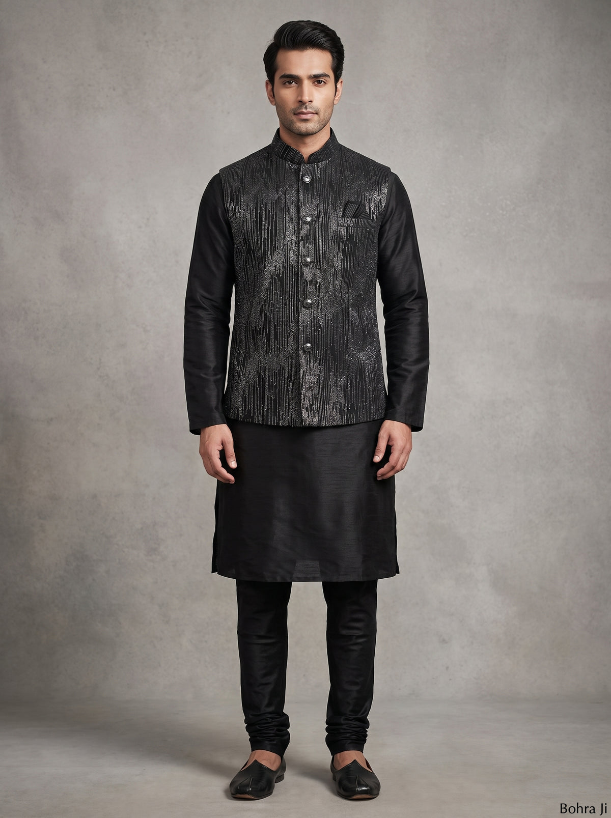 A man modeling a black traditional Indian outfit consisting of a textured black sherwani jacket, black kurta, black trousers, and black loafers, standing against a textured gray studio backdrop.