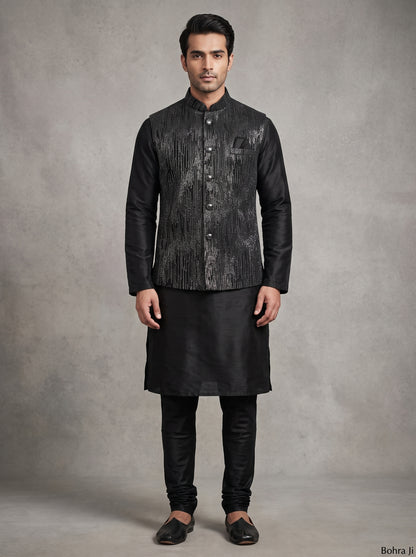 A man modeling a black traditional Indian outfit consisting of a textured black sherwani jacket, black kurta, black trousers, and black loafers, standing against a textured gray studio backdrop.