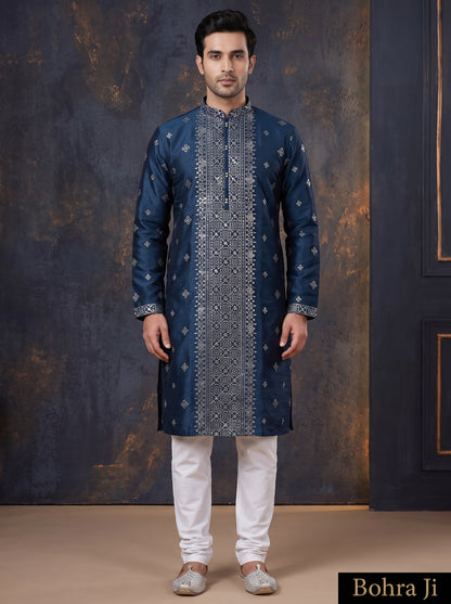 A man modeling a dark blue long kurta with intricate silver embroidery, paired with white trousers and silver shoes, standing against a dark textured studio backdrop. The brand name 'Bohra Ji' is visible in a black box in the bottom right corner.