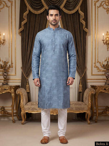 A man modeling a light blue patterned kurta with white trousers and brown shoes, standing in an ornate, luxurious room with gold furniture and brown drapes. The brand name 'Bohra Ji' is visible in a black box in the bottom right corner.
