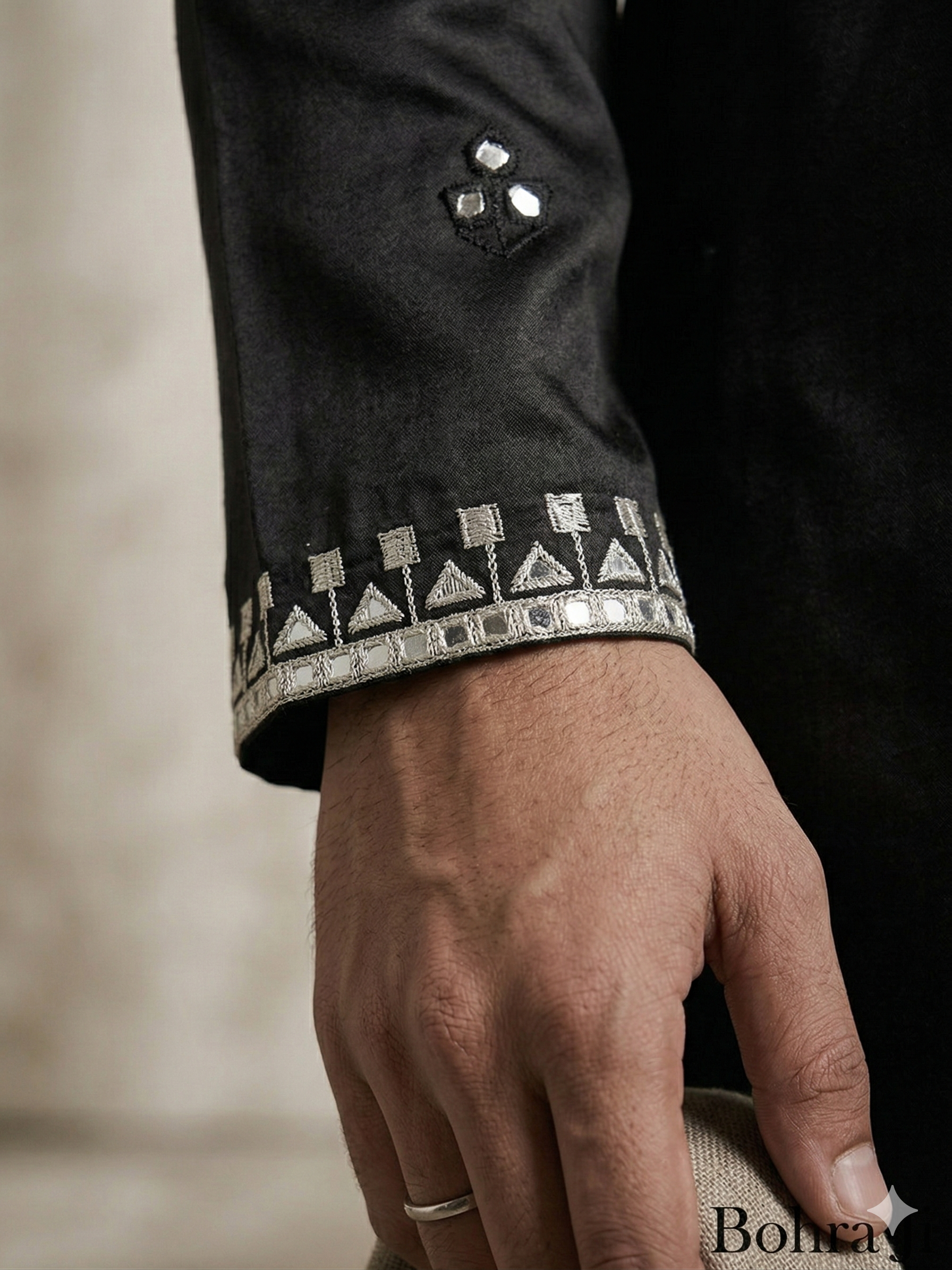 Close-up of a hand wearing a ring on a dark garment with intricate patterns.